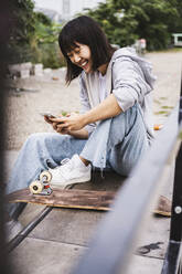 Fröhliches Teenager-Mädchen, das auf einer Bank sitzend ein Smartphone benutzt - UUF24749