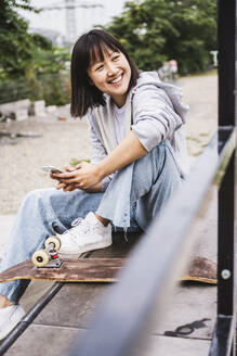Glücklicher weiblicher Teenager mit Smartphone auf einer Bank sitzend - UUF24750