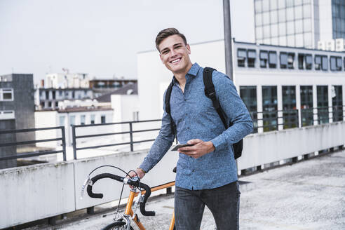 Lächelnder Mann mit Mobiltelefon und Fahrrad auf einem Hausdach - UUF24766