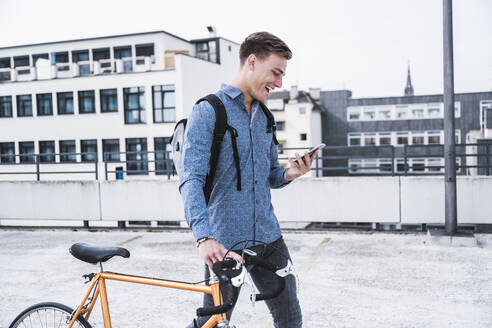 Fröhlicher Mann mit Fahrrad, der beim Spaziergang auf dem Dach eines Parkhauses ein Mobiltelefon benutzt - UUF24771