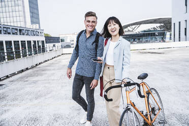 Happy male and female friends with bicycle walking on parking garage rooftop - UUF24774