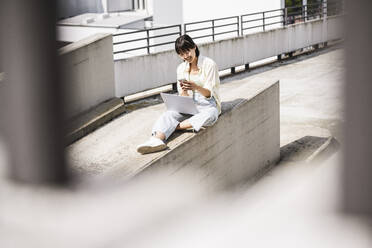 Teenage girl with laptop using smart phone on retaining wall during sunny day - UUF24807