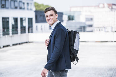 Smiling businessman with backpack walking on rooftop - UUF24820