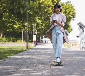 Lächelndes Teenager-Mädchen, das ein Smartphone benutzt, während es auf dem Gehweg Skateboard fährt - UUF24833