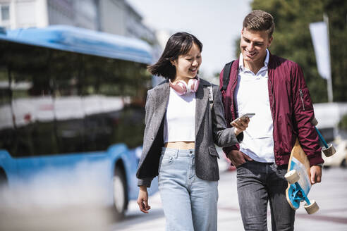 Lächelnde männliche und weibliche Freunde teilen sich ein Mobiltelefon während eines Spaziergangs an einem sonnigen Tag - UUF24837