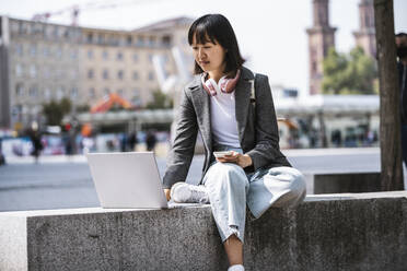 Geschäftsfrau, die einen Laptop benutzt, während sie auf einer Stützmauer sitzt - UUF24843