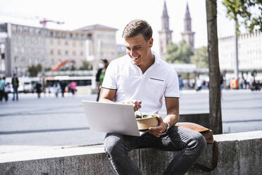 Lächelnder Mann mit Laptop beim Essen in der Stadt - UUF24846