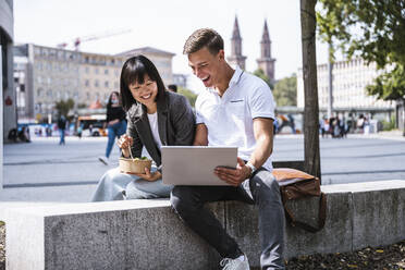 Happy male and female friends using laptop while sitting on retaining wall - UUF24847