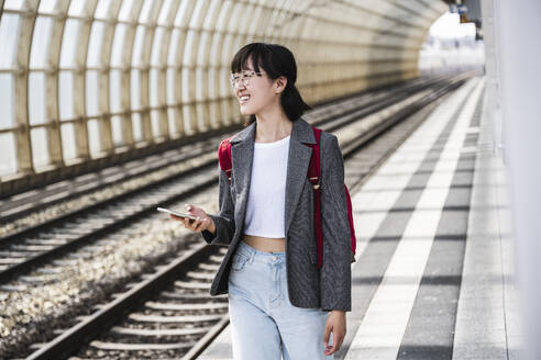 Lächelnder weiblicher Teenager mit Mobiltelefon am Bahnhof - UUF24850