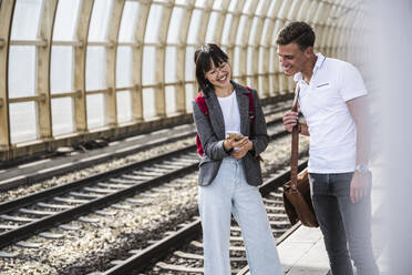 Glückliche männliche und weibliche Freunde teilen sich ein Mobiltelefon auf dem Bahnsteig - UUF24852