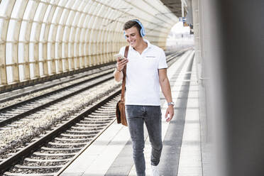 Young man using mobile phone while walking on train station platform - UUF24854