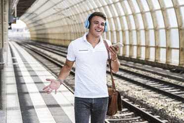 Young man wearing wireless headphones talking through mobile phone at train station - UUF24855