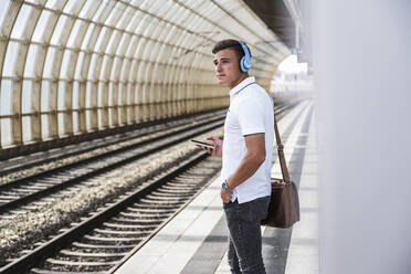Young man with mobile phone and shoulder bag standing at train station - UUF24856