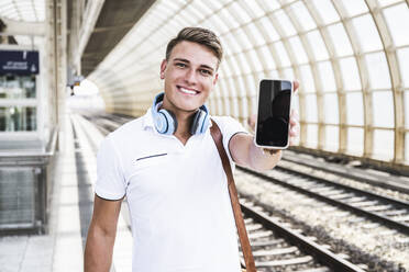 Lächelnder Mann zeigt Smartphone am Bahnhof - UUF24857