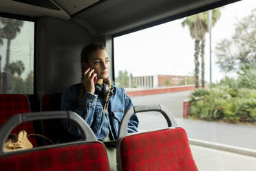 Junge Frau, die mit ihrem Handy telefoniert und durch das Busfenster schaut - VABF04403