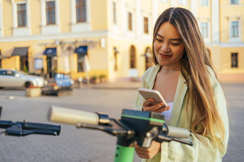 Frau mit Elektroroller in der Stadt, die ein Mobiltelefon benutzt - OYF00520