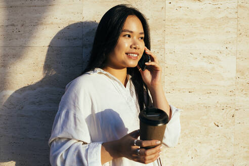 Glückliche Frau mit Einweg-Kaffeebecher im Gespräch mit dem Handy an einem sonnigen Tag - OYF00529