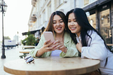 Female friends sharing mobile phone at sidewalk cafe - OYF00541