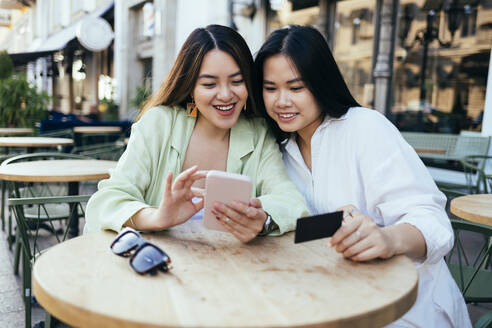 Smiling women shopping online through smart phone at sidewalk cafe - OYF00542