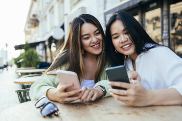 Female friends using mobile phones while sitting at sidewalk cafe - OYF00546