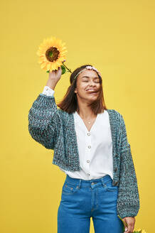 Glückliche junge Frau mit Sonnenblume vor einer gelben Wand - KIJF04135