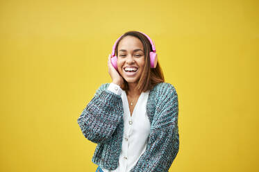 Happy woman listening music through wireless headphones in front of yellow wall - KIJF04150
