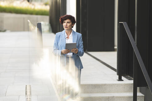 Mature businesswoman with digital tablet standing on staircase outside office building - JCCMF04059