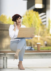 Mature female professional talking on smart phone while sitting with laptop at office park - JCCMF04066