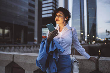 Beautiful businesswoman holding mobile phone while standing by railing of bridge during dusk - JCCMF04093