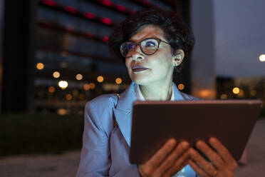 Schöne reife Geschäftsfrau mit Brille hält digitales Tablet in der Nacht - JCCMF04099