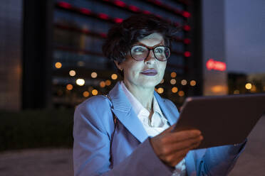 Reife weibliche Fachkraft mit Brille, die nachts ein digitales Tablet hält - JCCMF04100