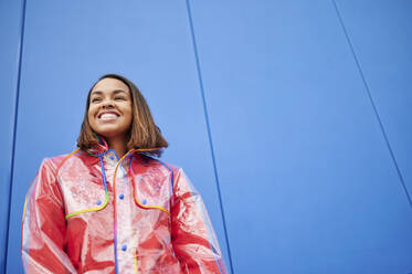 Smiling woman wearing raincoat in front of blue wall - KIJF04153