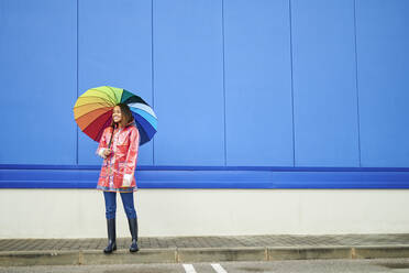 Frau mit mehrfarbigem Regenschirm auf dem Fußweg an der blauen Wand stehend - KIJF04156
