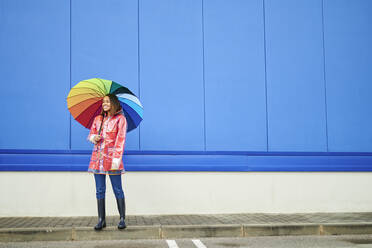 Woman with multi colored umbrella standing on footpath by blue wall - KIJF04156