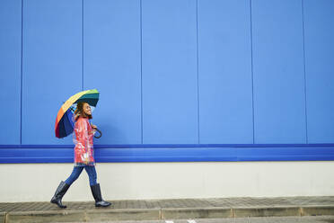 Woman wearing raincoat holding umbrella while walking on footpath - KIJF04157