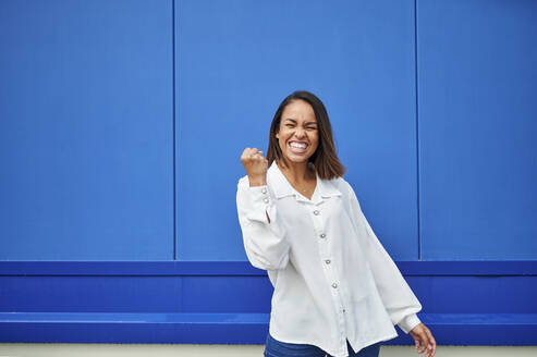 Happy young woman cheering in front of blue wall - KIJF04170