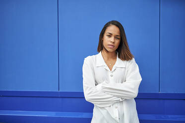 Young woman with arms crossed standing in front of blue wall - KIJF04171