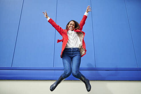 Carefree woman with hands raised jumping in front of blue wall - KIJF04172