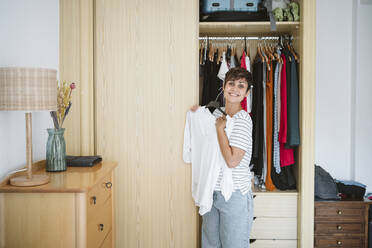 Smiling woman holding dress near closet at home - EBBF04591