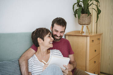 Happy couple with notebook sitting on bed at home - EBBF04611