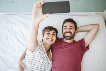 Frau nimmt Selfie mit Mann durch Smartphone, während auf dem Bett liegend zu Hause - EBBF04616