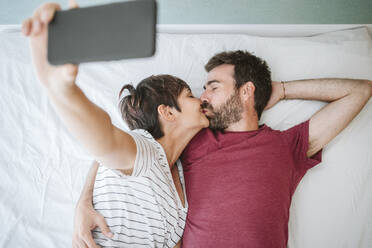 Affectionate couple kissing while taking selfie on bed at home - EBBF04617