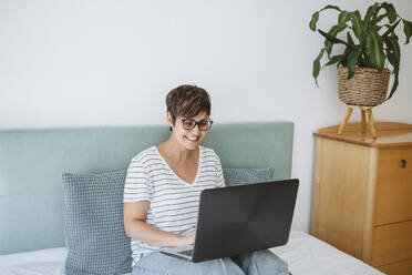 Smiling woman using laptop on bed at home - EBBF04622