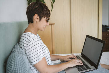 Mittlere erwachsene Frau, die einen Laptop benutzt, während sie im Schlafzimmer sitzt - EBBF04631