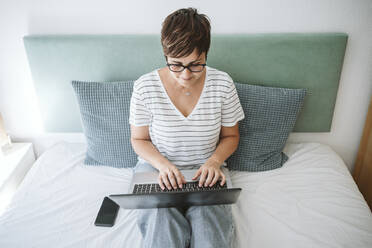Frau mit kurzen Haaren benutzt einen Laptop, während sie zu Hause auf dem Bett sitzt - EBBF04632