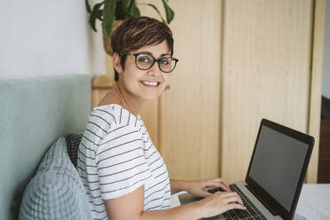 Lächelnde Frau mit Brille sitzt mit Laptop zu Hause - EBBF04633