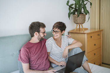 Lächelndes erwachsenes Paar mit Laptop, das zu Hause auf dem Bett sitzt - EBBF04636