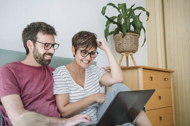 Lächelndes mittleres erwachsenes Paar, das einen Laptop benutzt, während es auf dem Bett sitzt - EBBF04638