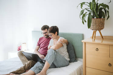 Man with woman using laptop while sitting on bed at home - EBBF04639