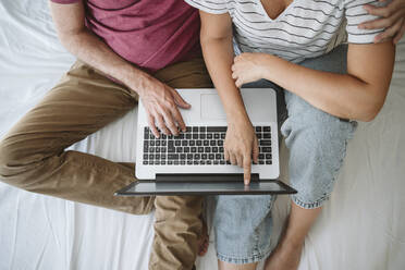 Frau berührt den Bildschirm, während sie mit einem Mann auf dem Bett sitzt und einen Laptop benutzt - EBBF04640
